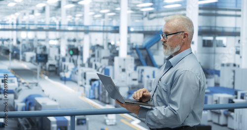 Portrait of a Handsome Middle Aged Engineer with Glasses Using Laptop Computer and Looking Around a Factory Facility with Equipment Producing Modern Electronic Components for Different Industries