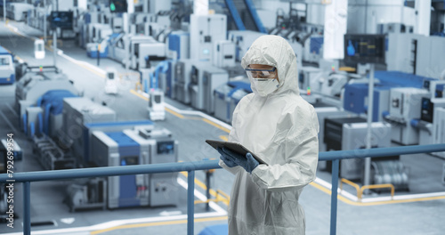 Female Technician in Protective Coveralls, Mask and Eyewear Using Tablet Computer at a Manufacturing Complex. Specialist Monitoring Conditions at a Modern Electronics Factory with Automated Robots © Gorodenkoff