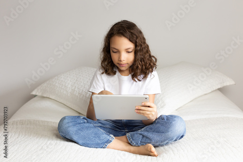 Cute mixed race nine-year old girl, sitting crossed-legs on bed holding a tablet and looking at its screen