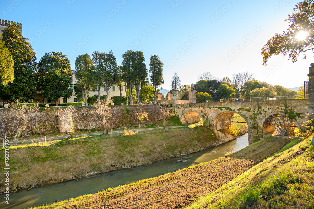 A narrow river flows under a stone medieval bridge against the backdrop of spring vegetation and blue sky