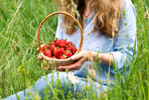 A young woman holds a basket of ripe strawberries while sitting on the grass. Healthy eating, outdoor recreation. Copy space. Selective focus.