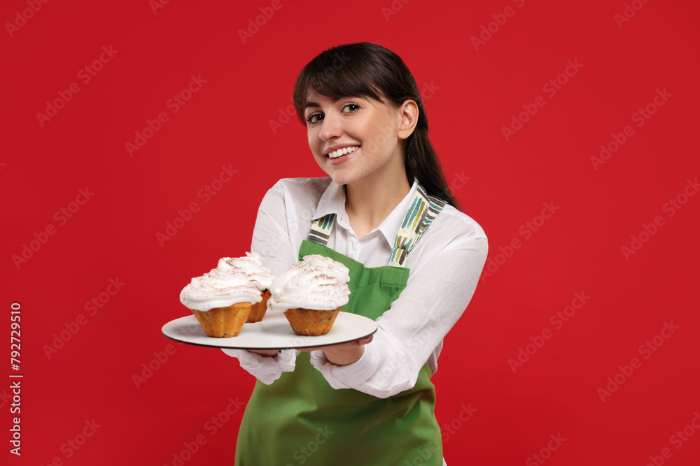 Happy professional confectioner in apron holding delicious cupcakes on red background Happy professional confectioner in apron holding delicious cupcakes on red background