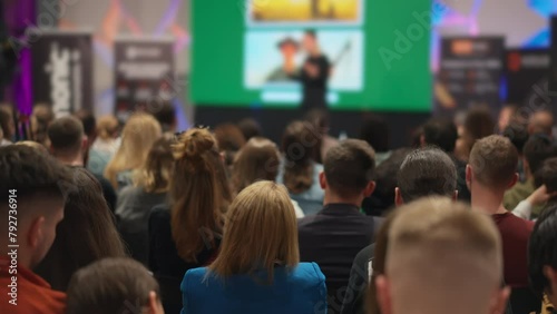 Young audience engaged at entrepreneurship conference, female speaker presents on stage. Diverse group of men and women focused on business incubator event. Concept: entrepreneurship and startup