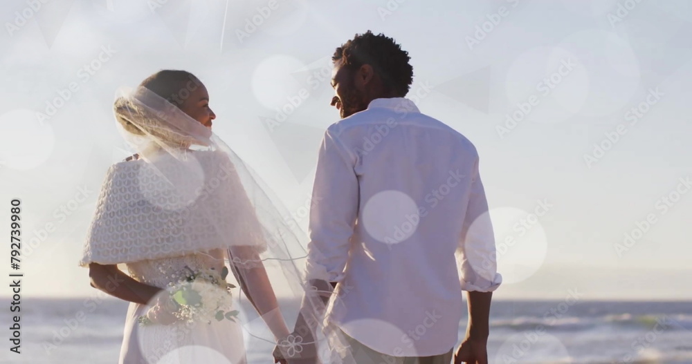 Naklejka premium Image of light spots over african american bride and groom holding hands on beach at wedding