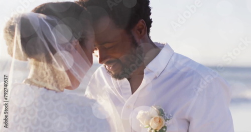Image of light spots over happy african american groom unveiling bride on beach at wedding