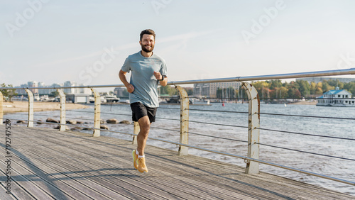 Fototapeta Naklejka Na Ścianę i Meble -  A joyful jogger with a light beard advances along a wooden pier by the river, cityscape and boats in the soft-focus background.