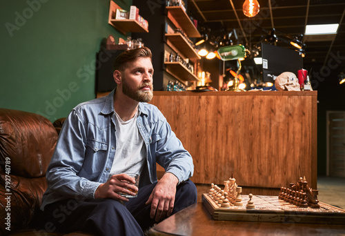 Handsome man resting after appointment in modern barbershop. Male client drinking whiskey. Bearded man sitting on leather couch in front of chessboard.