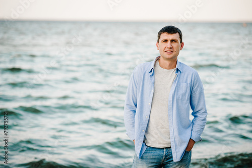Handsome man joyfully walks near sea and looks at camera. Young traveler man standing on beach ocean and enjoying summer day on vacation. Portrait of stylish male enjoying amazing view.