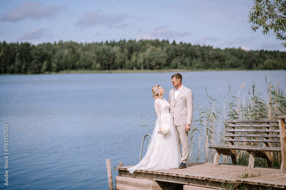 Valmiera, Latvia - August 10, 2023 - Bridal couple standing on a dock by a serene lake, surrounded by forest, exchanging a loving glance...