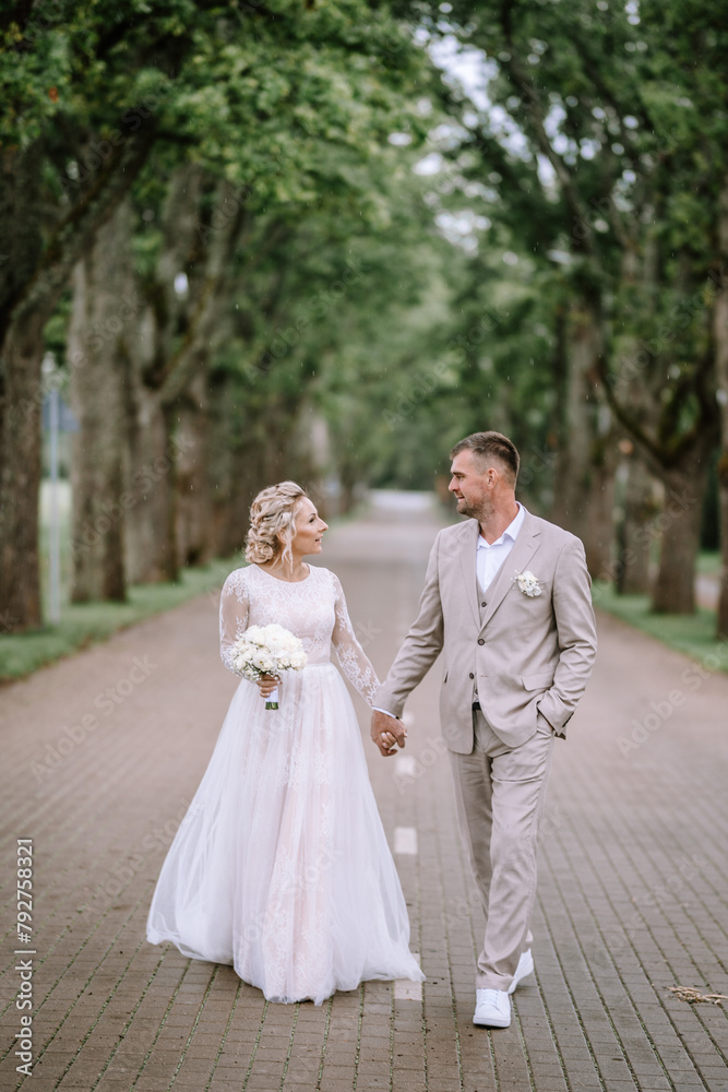 Valmiera, Latvia - August 10, 2023 - A bride and groom walking hand in hand on a cobblestone path lined with tall trees, exchanging smiles. Valmiera, Latvia - August 10, 2023 - A bride and groom walking hand in hand on a cobblestone path lined with tall trees, exchanging smiles.