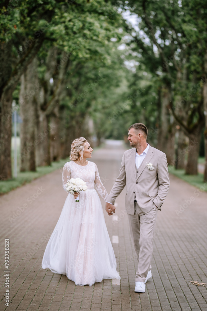 Valmiera, Latvia - August 10, 2023 - A bride and groom walking hand in hand on a cobblestone path lined with tall trees, exchanging smiles.