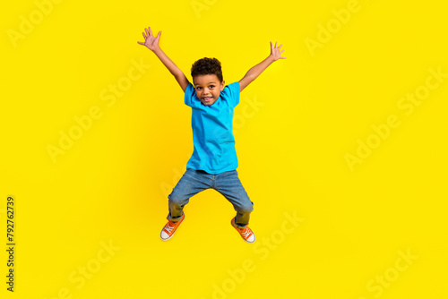 Full size photo of overjoyed little schoolboy wear blue t-shirt denim pants raising palms jumping isolated on vivid yellow background © deagreez