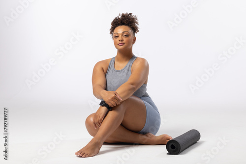 Biracial young female plus size model sits next to yoga mat on a white background, copy space
