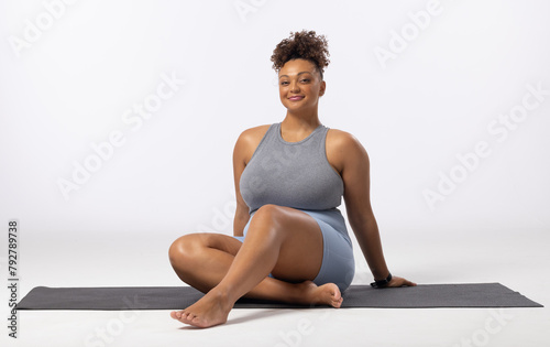 A biracial young female plus size model sits on yoga mat, smiling, copy space
