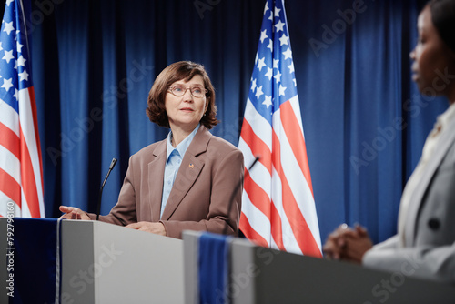 Confident female politician in eyeglasses and formalwear speaking to opponent or foreign colleague while standing by platform