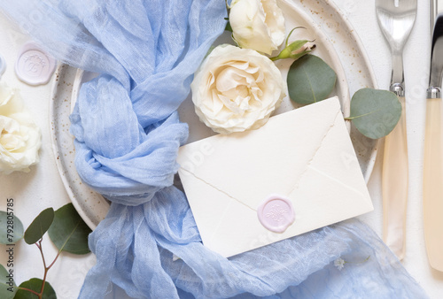 Envelope near blue tulle fabric knot and cream roses on plates top view copy space, wedding mockup