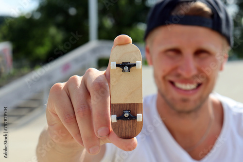 Happy young smile man holding blank wooden fingerboard in skatepark. Mini skateboard, small skateboard deck