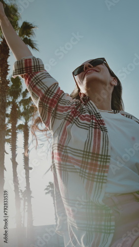 Cheerful young woman in sunglasses raises hand leaning out of car window against tall palm trees