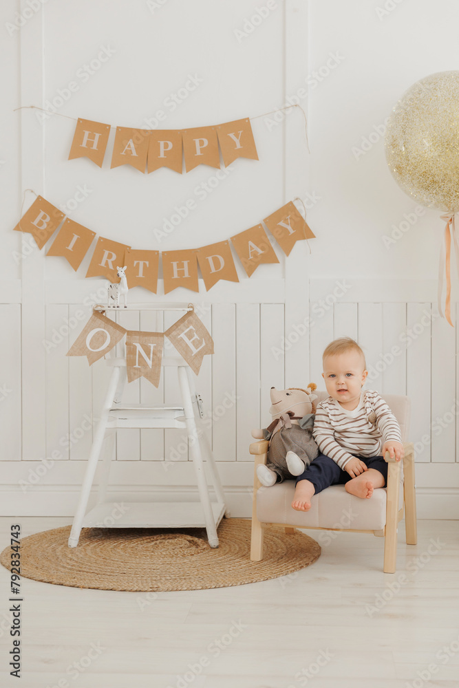 Naklejka premium Baby first birthday concept. Portrait Happy caucasian little boy sits against background of holiday decorations and flags with inscription.