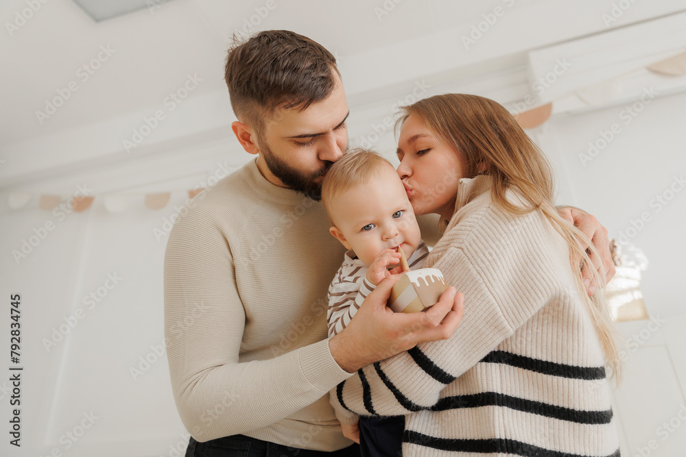 Fototapeta premium Happy baby boy receives kisses from his loving parents, creating heartwarming scene during first birthday celebration at home. Lifestyle moment of scandinavian family