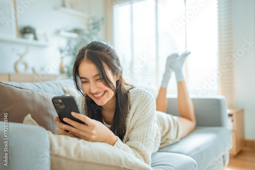 Happy young asian woman lady browsing surfing wireless internet on mobile phone while sitting a sofa couch in living room at home apartment, Shopping online via website © oatawa