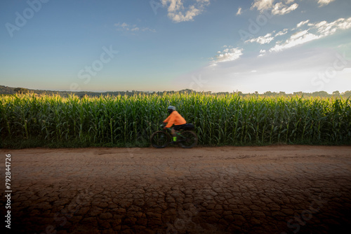 Ciclista de roupa laranja,  passando por Estrada rural, Plantação de Milho e céu azul