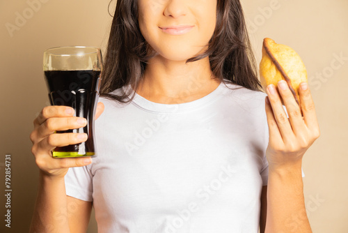 unrecognizable young woman dressed in a white shirt holding an empanada, salteña and a glass of cola soda
