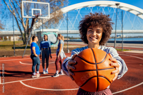 Portrait of young woman with her female friends on basketball court looking at camera and holding ball in her hands.