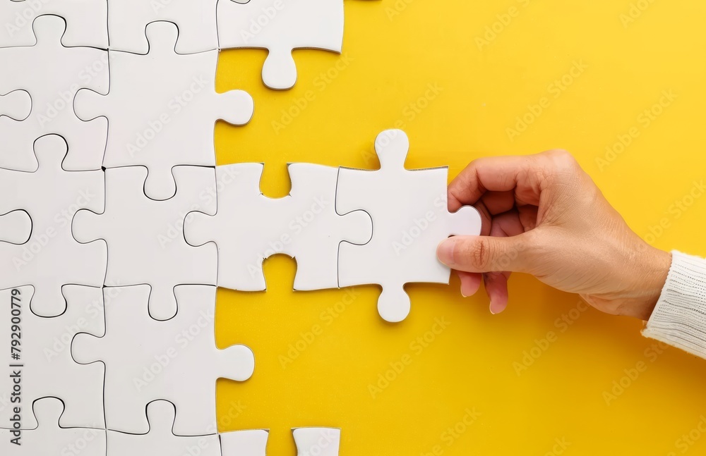 Obraz premium A person hand holding the last puzzle piece to complete a white puzzle on a yellow background