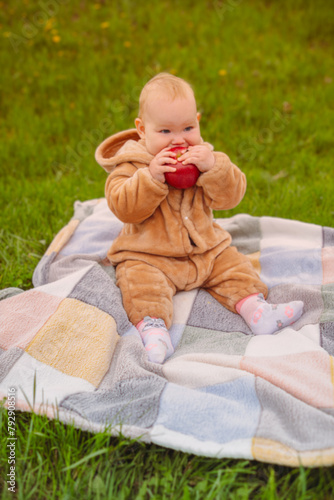 Cute baby girl sitting on plaid and eating apple on the green lawn