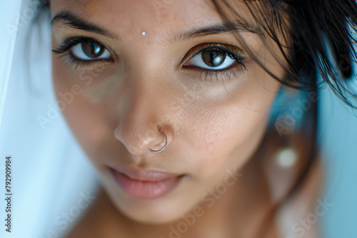 A young Indian woman with smooth, clean facial skin poses for a cropped portrait. With a nose stud and long eyelashes, she looks directly at the camera,  photo