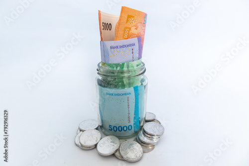Indonesian Rupiah banknotes in glass jar and scattered coins on white background