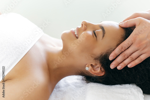 Side-view on calm relaxed smiling brazilian or hispanic woman lies on a massage table in a salon during a forehead massage. Female massage therapist doing general forehead massage to a pretty girl