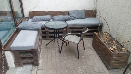 Metal chairs and a table with a glass top stand on a tile platform. Nearby, near the concrete walls, pallet seats with mattresses and cushions are made and a wooden flower bed is set up