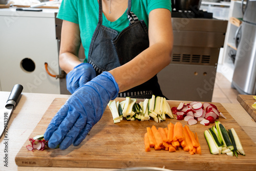 Unrecognisable woman cutting vegetables on a board, carrots, radish, cucumber to go with hummus. Detail of hands with blue latex gloves.