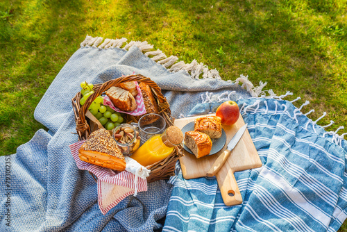 Picnic duvet and basket with different food, fruits, orange juice., yogurt and bread on green grass
