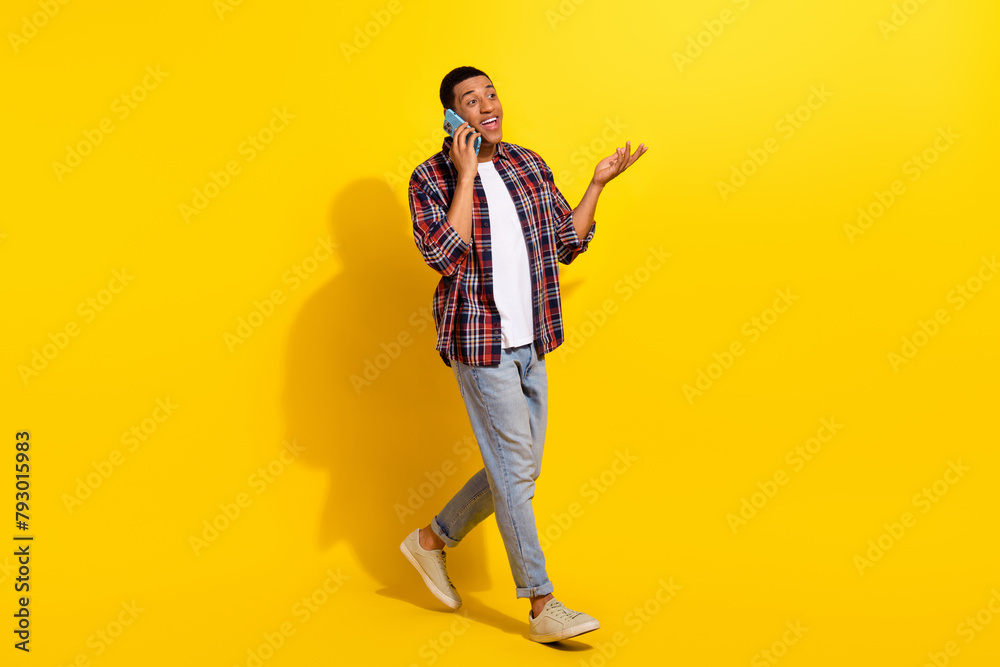 Full length photo of smart guy wear checkered shirt denim pants walk in empty space talk on smartphone isolated on vivid color background Full length photo of smart guy wear checkered shirt denim pants walk in empty space talk on smartphone isolated on vivid color background