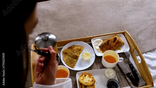 Woman is eating desserts in bed from wooden tray in hotel room on vacations, top view. Pancakes, cheesecakes, pastry with tea and ice-cream, condensed milk, sour cream and jam. Resting and relax.