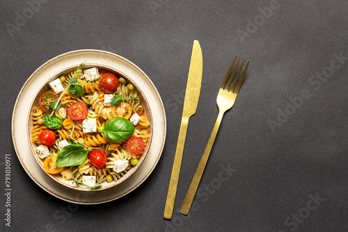 Colorful full grain fusilli pasta warm salad with feta cheese, cherry tomatoes, herbs, green pea and basil leaves on black stone background top view, copy space for your design.