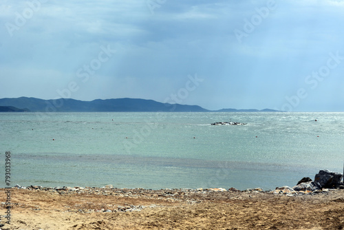 Beach shore with turquoise sea and distant mountains under blue sky.
