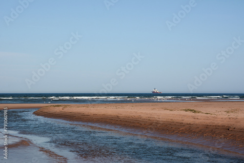 A wide beach with shallow smooth sand. In the distance, a ship sails in the sea under a blue sky