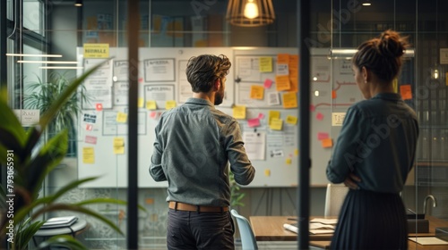 In the image  two people are standing in front of a whiteboard with sticky notes. The whiteboard is filled with notes  and the people seem to be observing or discussing them.