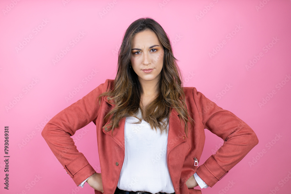 Young beautiful woman wearing casual jacket over isolated pink background skeptic and nervous, disapproving expression on face with arms in waist Young beautiful woman wearing casual jacket over isolated pink background skeptic and nervous, disapproving expression on face with arms in waist