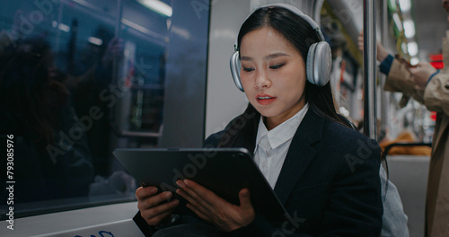 Beautiful Asian woman sitting next to window while watching video or movie on large tablet device. Listening to music in headphones. Tapping on screen and beginning to smile. Public transportation.