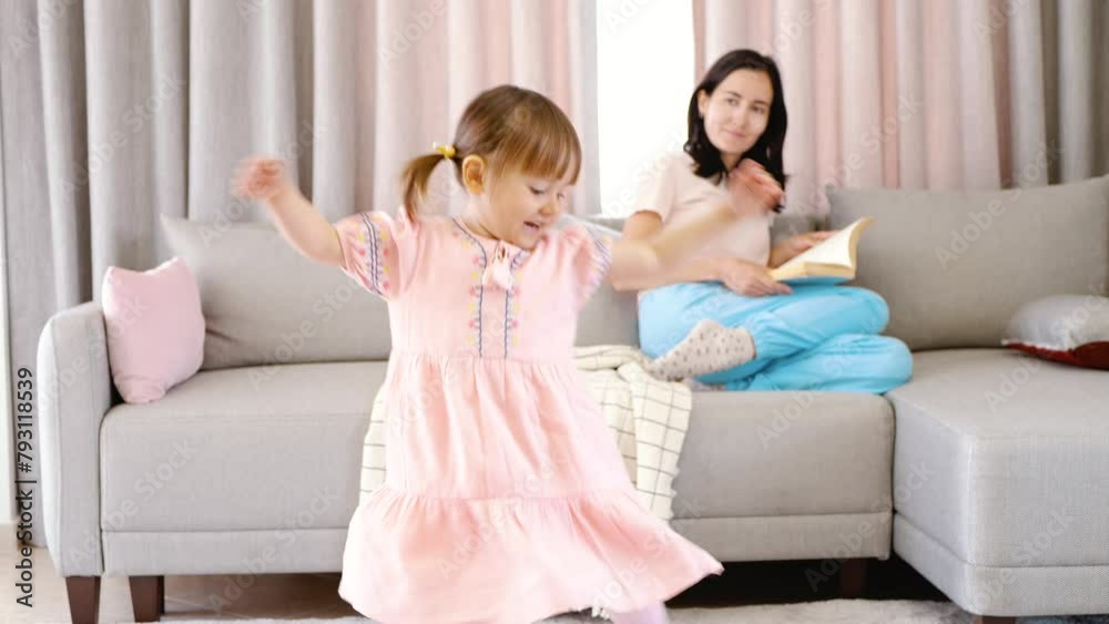 The child is dancing in the room. In the background, mom is reading a book on the sofa. The child is dancing in the room. In the background, mom is reading a book on the sofa.