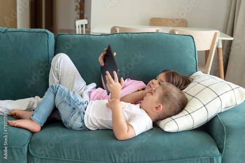 kids lie on the sofa in the living room, looking at the tablet. Making parent call, playing video game, watching online content, browsing internet