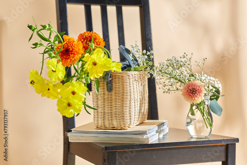 home decor and design concept - close up of flowers in basket and magazines on vintage chair over beige background