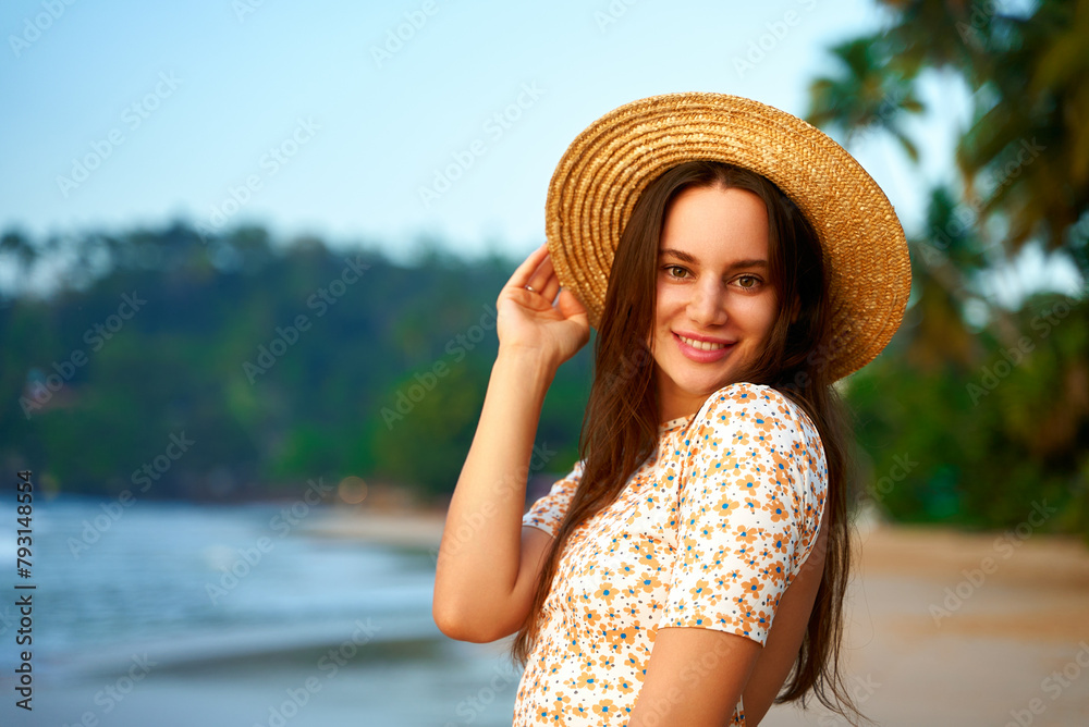 Smiling woman in floral dress and straw hat enjoys tropical beach at sunset. Summer fashion, leisure by sea, female traveler explores coast, poses with ocean backdrop, retro vibe in holiday attire.