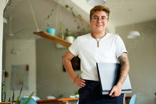 Transgender professional in bright office smiles, holding laptop, inked arms visible. Confident stance inclusive work culture advocate. Nerdy glasses smart casual work attire. Plants modern workspace.