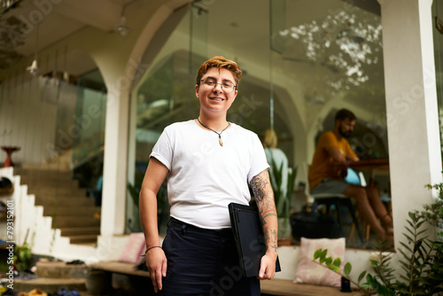 Smiling transgender pro holds laptop in modern workspace. Inclusive work environment with diverse colleagues. Casual business attire, gender diversity, trans male at creative office.